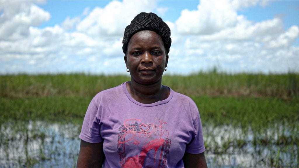 Angelina, een vrouw uit Mozambique, kijkt in de camera. Op de achtergrond een overstroomd grasveld.