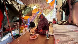 Een zwangere vrouw zit midden in een zelfgemaakte tent met een meisje in de achtergrond en een klein bord met fruit voor haar. Er ligt een matras naast haar met bloemetjespatroon.