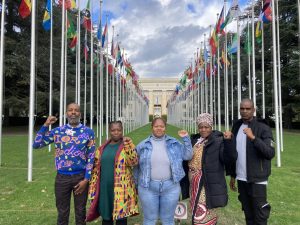 De 5 leden van MACUA/WAMUA staan buiten in Geneve met de vlaggen en het VN-gebouw in de achtergrond.