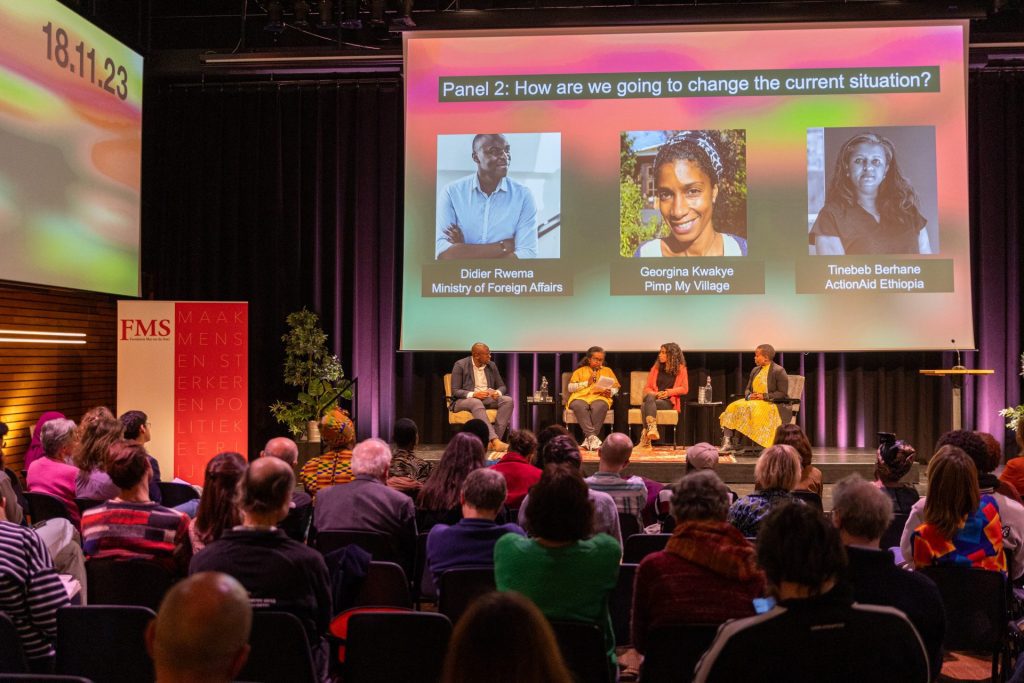 Tinebeb Berhane aan het woord op het podium van Pakhuis de Zwijger. Je ziet groot boven haar en haar mede panelleden hun naam en foto op het scherm. Links van Tinebeb zit Didier Rwema, van Buitenlandse Zaken. Rechts van haar zit Georgina Kwakye, van Pimp My Village. En verder rechts zit de moderator (en geweldig afdelingshoofd van ActionAid Nederland) Sophie Kwizera in een prachtige gele jurk. Vooraan de foto zie je een volle zaal met mensen die aandachtig luisteren naar Tinebeb.