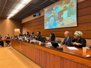 Een rij van 8 mensen zit achter een lange tafel met microfoons voor zich, waarbij WAMUA-lid Khosi het dichtst vooraan zit. Op een beeldscherm boven ze is een vrouw te zien die een vraag aan het stellen is.