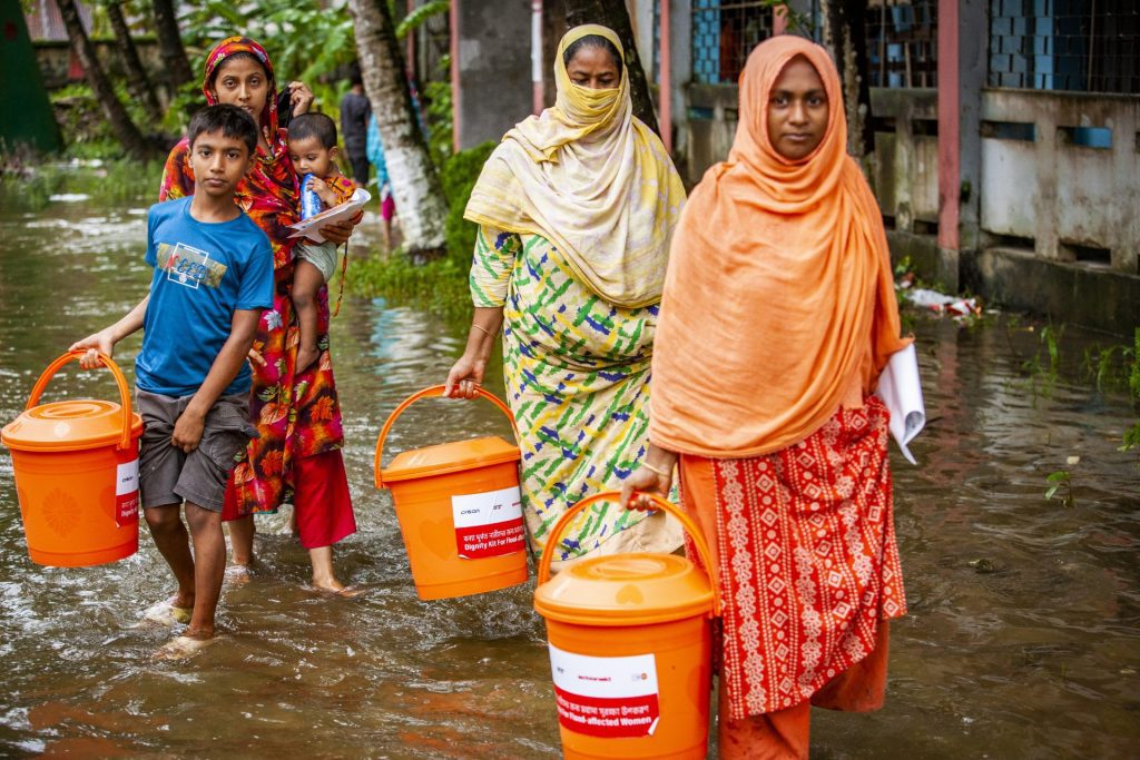 Vrouwen lopen door het water met oranje emmers vol noodspullen van ActionAid.
